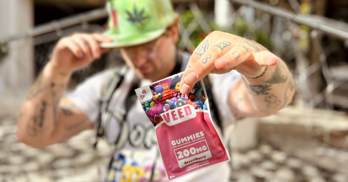 Man wearing a green cannabis hat holding a VEED 200mg THC gummies package outdoors.